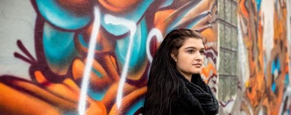 Young person in scarf stood in front of graffiti'd wall.